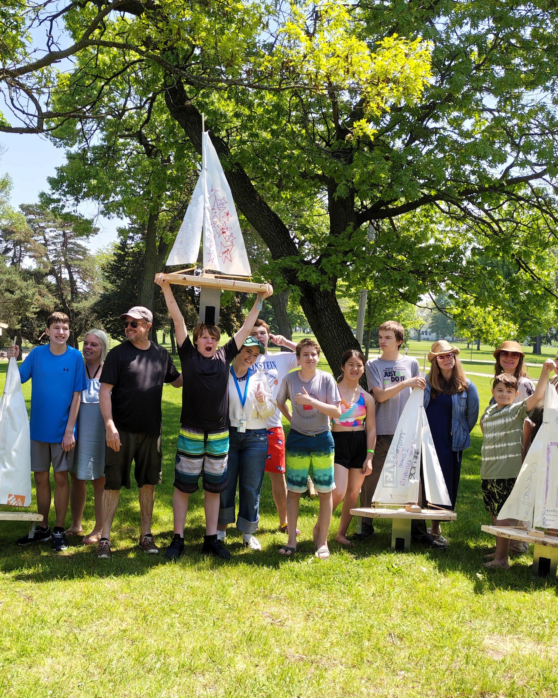 Students, staff and families gathered under trees in a sunny park, holding the model sailboats they built.