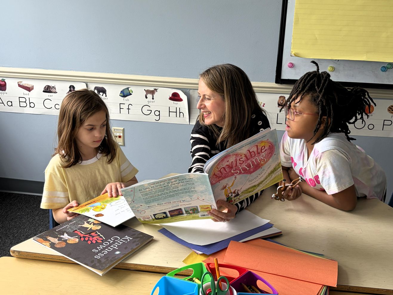 A teacher reads a picture book with two students at a classroom table, an alphabet chart on the wall behind them.