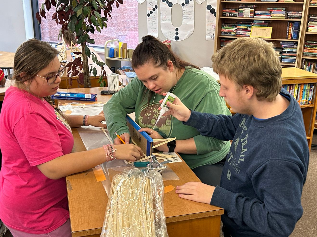 A teacher and two students collaborate on a hands-on building project at a classroom table, with bookshelves and student work in the background.