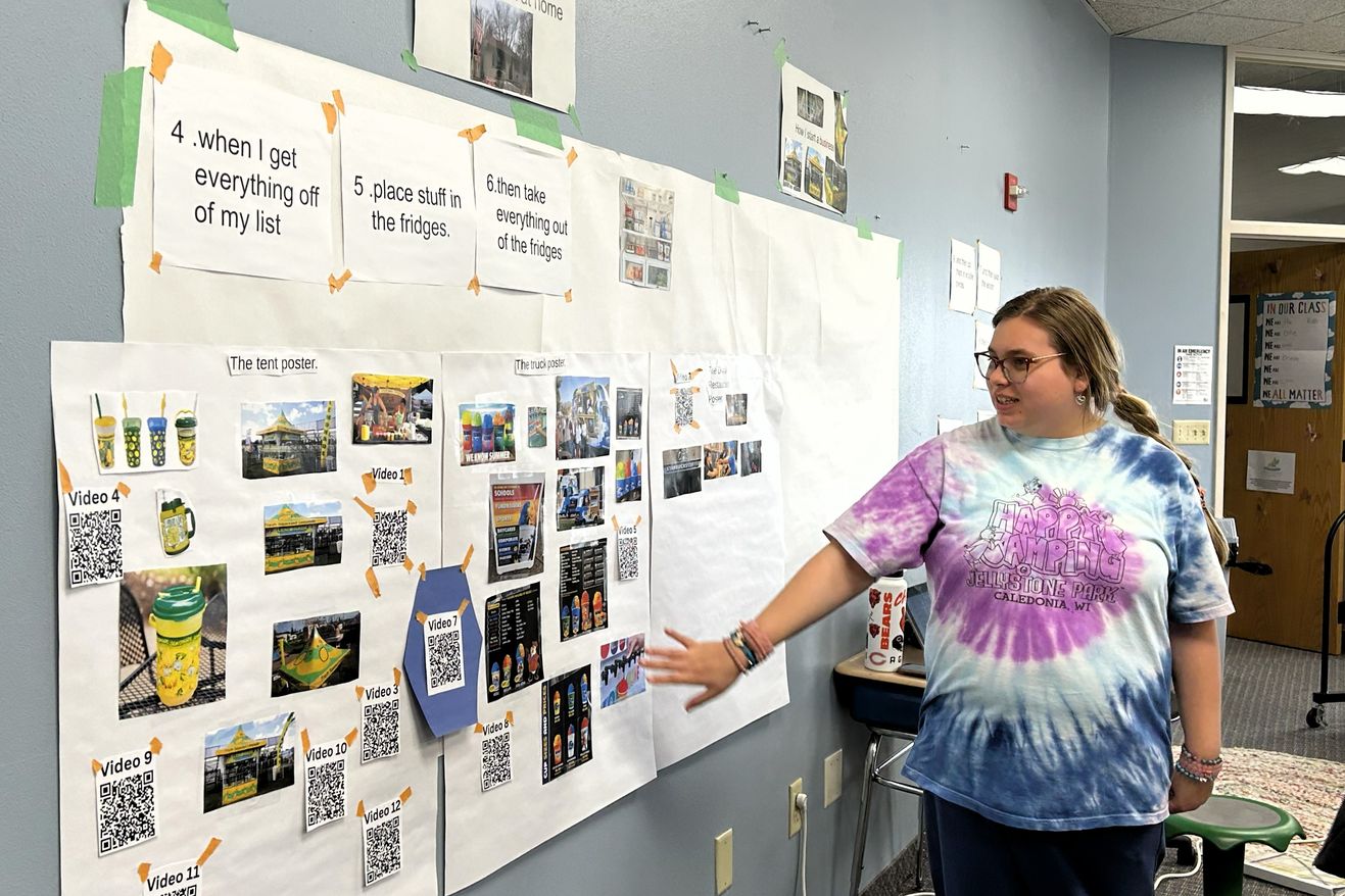 A student presents a research project with photos and QR codes displayed on the classroom wall.