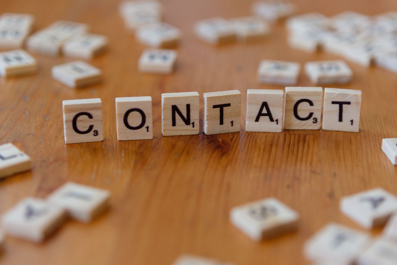 Wooden Scrabble letter tiles spelling out the word CONTACT on a table, with scattered tiles around them.
