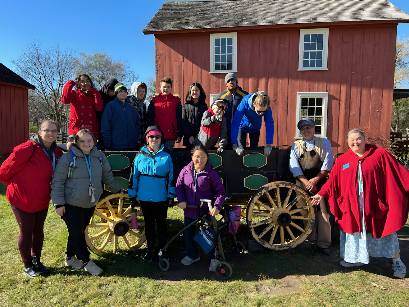 Students and staff pose together on an old wooden wagon in front of a red barn during a field experience at a historic farm.