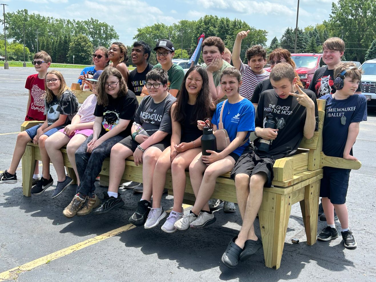 A large group of Open Wings Upper School students and staff packed together on a wooden bench outdoors, smiling on a summer day.