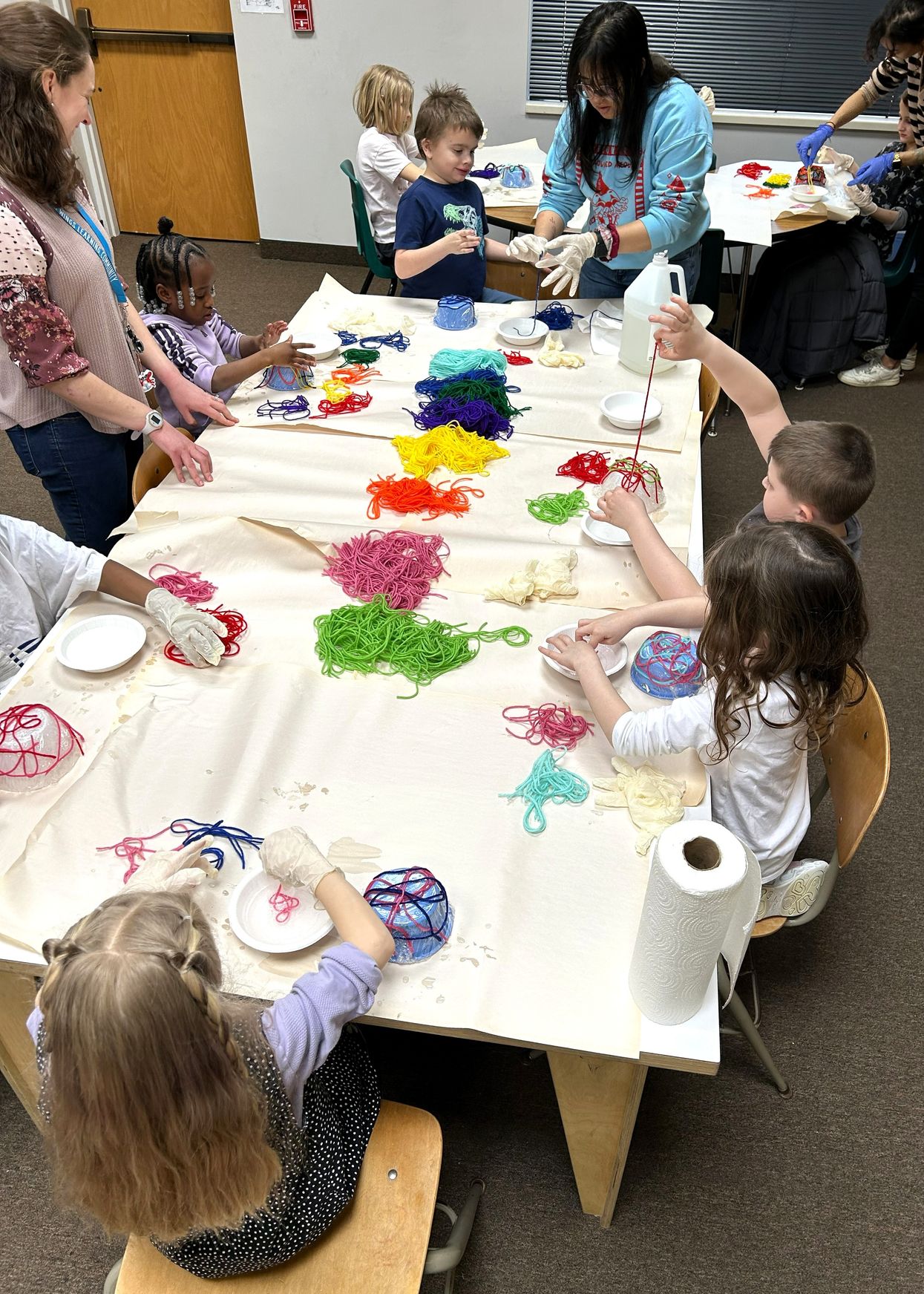 Students and staff around a long table working on a yarn and glue art project, with brightly coloured yarn spread across the paper.
