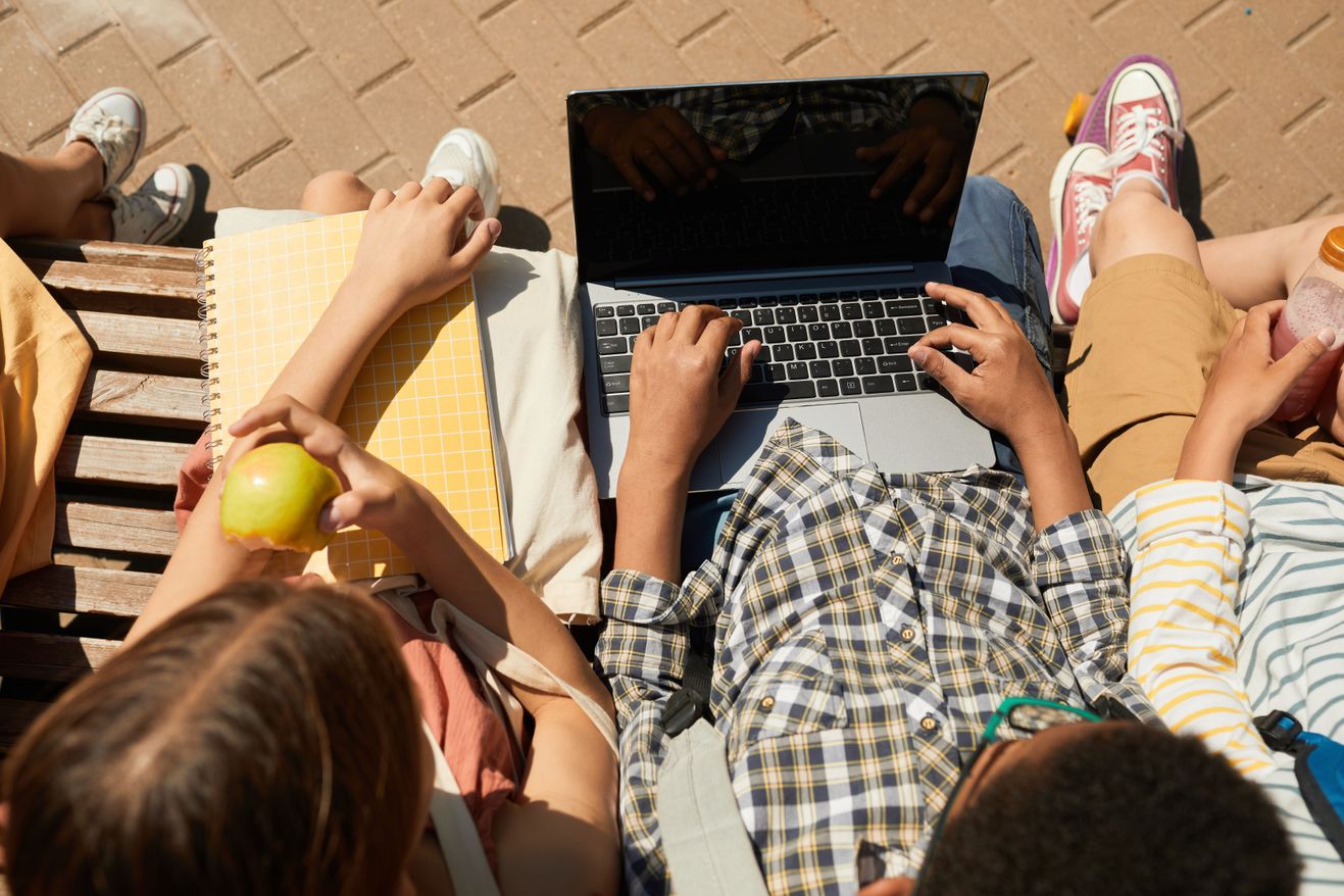 Students sitting together outdoors on a bench, one working on a laptop while others talk and share snacks.