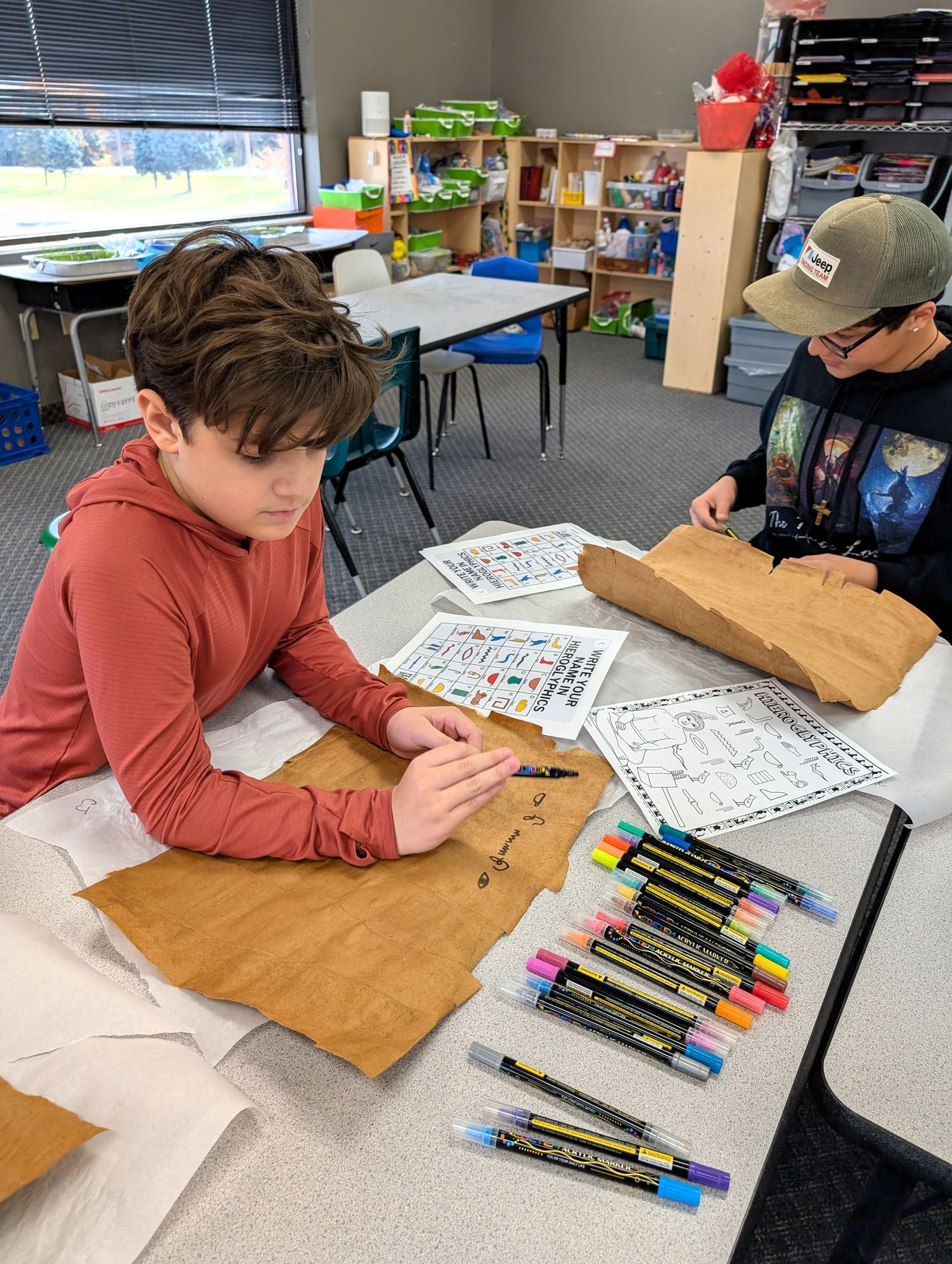 Two students write their names in hieroglyphics on faux parchment with coloured markers, using a hieroglyph key sheet.
