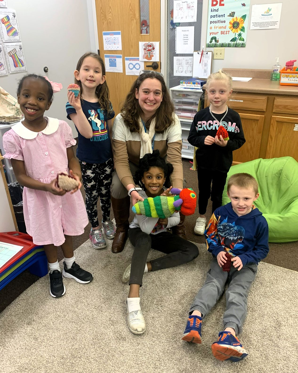 A teacher sits on the classroom floor with five young students, each holding a crocheted animal or treat they made.